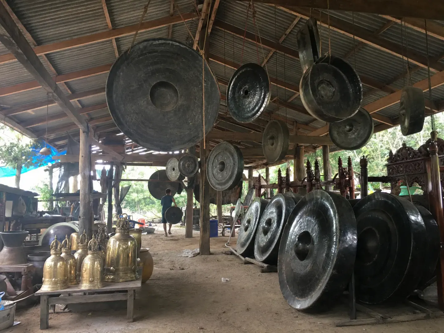 An open-air workshop with many finished gongs hanging overhead and large dark gongs below
